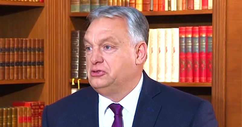 Orbán Viktor sötétkék zakóban, fehér ingben, lila nyakkendőben, ősz hajjal , furcsa fejjel beszél. Mögötte könyvekkel megrakott könyvespolc látható.