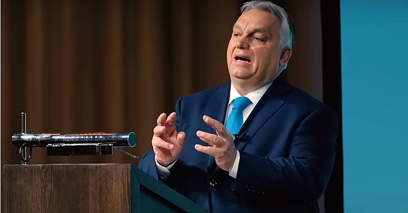 Orbán Viktor sötétkék zakóban, fehér ingben, világoskék nyakkendőben, ősz hajjal magyaráz a pódiumon, és közben hevesen gesztikulál.
