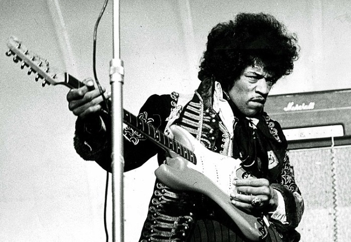 Jimi Hendrix rockzenészről egy fekete-fehér kép, amint gitározik egy koncerten. 
