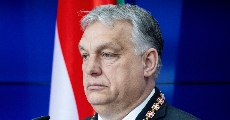 Orbán Viktor kimerülten balra nézve áll egy kék háttér előtt felállított magyar zászló előtt. A nyagában egy szerb zászlós nyaklánc (kitüntetés) van.