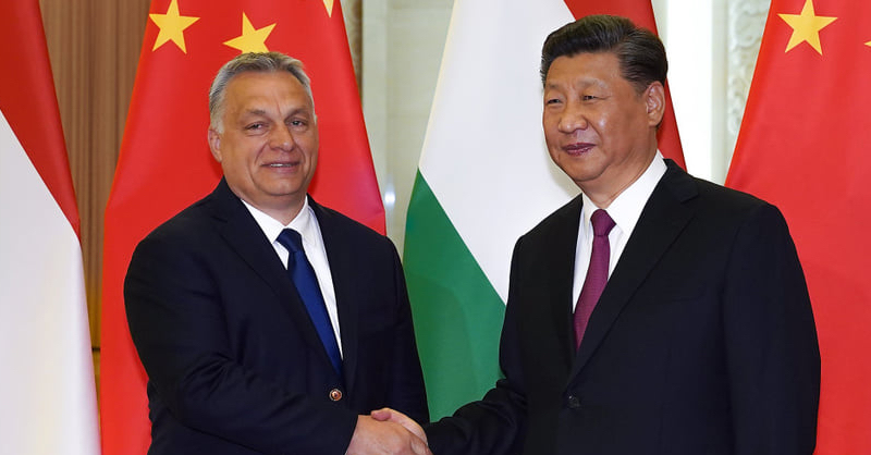 Orbán Viktor, Hszi Csin-Ping kezet fog sötét öltönyben, nyakkendőben, kínai és magyar zászlók előtt. Nagyon vigyorognak.